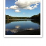 En blank sjö med speglingen av vita moln på en blå himmel.